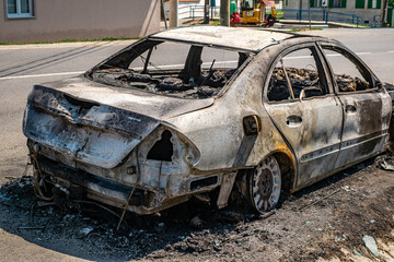 A color photograph captures a charred, severely damaged car sitting on a street's edge. The ground...