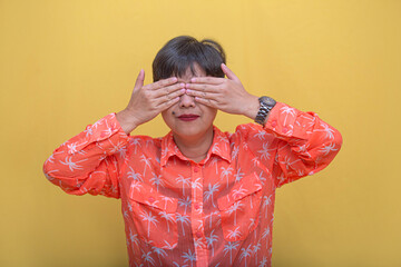 Portrait of a short haired Asian woman with gentle smile covering her eyes with two hands, making...