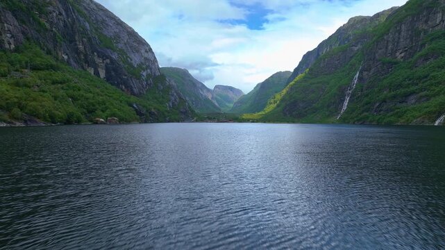 Aerial view of a fjord with steep green cliffs, a small settlement with red roofs by the water, and a drone moving forward over the tranquil scene.
