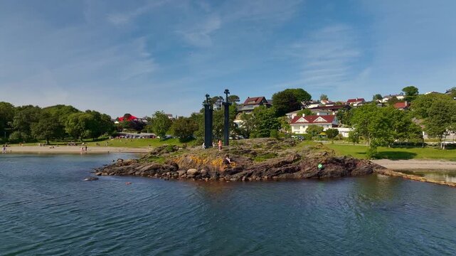 Drone captures Sverd i Fjell monument near Hafrsfjord, Norway, with bronze swords, green spaces, a beach, and hillside homes under clear skies.