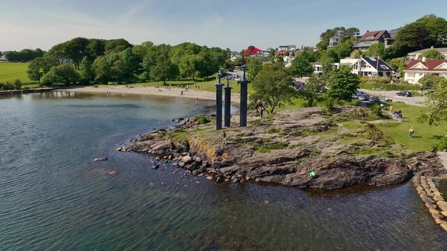 Aerial view of the Sverd i Fjell monument near Stavanger, Norway, with calm water, a small beach, lush greenery, and a residential neighborhood.