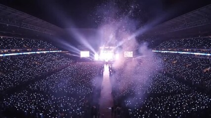 A massive stadium concert features a vibrant stage show with pyrotechnics, illuminating a sea of cheering fans. - Powered by Adobe
