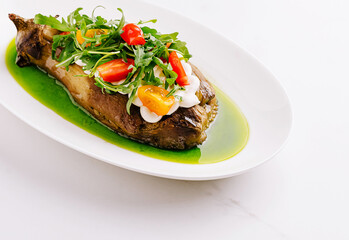 Roasted eggplant with cream, arugula, and cherry tomatoes, drizzled with a bright green sauce, served on a white plate.