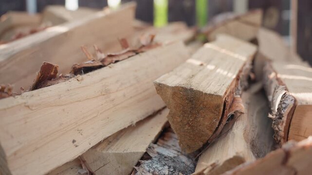 Close up of neatly stacked split logs resting on paved ground outdoors near wood fence under soft blur background and warm natural light highlighting wood grain textures, bark edges