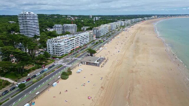 Drone reversing shot of La Baule-Escoublac beach, showing coastal road, buildings, surrounding trees, and beach activity with people walking and sunbathing under umbrellas - France