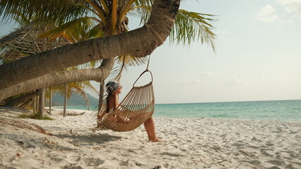 Woman relaxing in a hanging hammock chair under palm trees on a sandy tropical beach, enjoying...