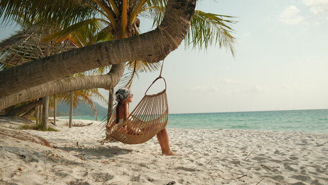 Woman relaxing in a hanging hammock chair under palm trees on a sandy tropical beach, enjoying peaceful seaside vacation and summer lifestyle.
