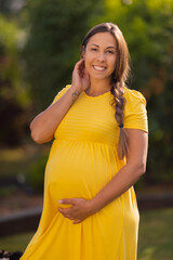 Smiling pregnant woman in yellow dress touching her belly outdoors