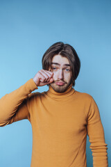 Fototapeta premium portrait of young man in a turtleneck displays sadness, actively wiping his eye as a sign of distress on blue background