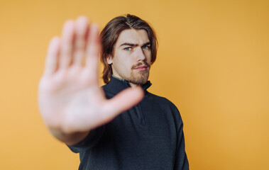 portrait of young man holds his hand up, looking serious as if signaling to pause on yellow...