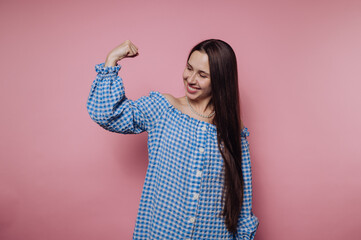 portrait of young woman flexes her bicep proudly while smiling in a blue checkered outfit on pink...