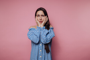 portrait of young young woman expresses concern and worry while touching her face on pink background