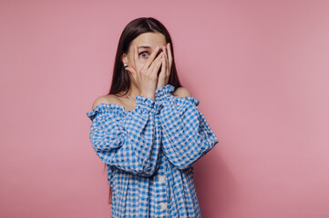 portrait of young woman playfully hides her face with her hands, expressing surprise on pink...