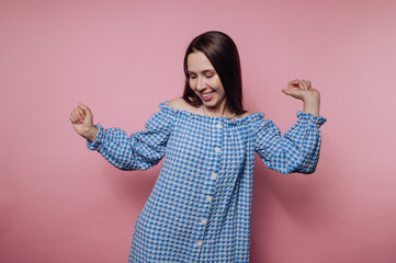 portrait of young woman joyfully dances while wearing a blue checkered dress on pink background
