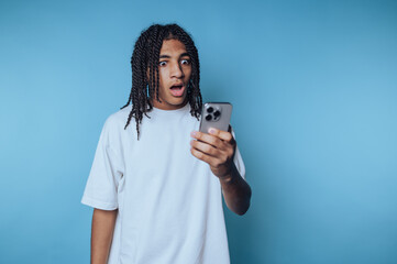 portrait of young man gazes at their smartphone, clearly shocked on blue background