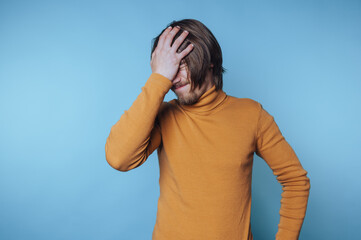 portrait of young man in a turtleneck shows frustration with a facepalm gesture on blue background