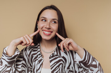portrait of young playful woman gives a bright smile while resting her fingers on her cheeks,...