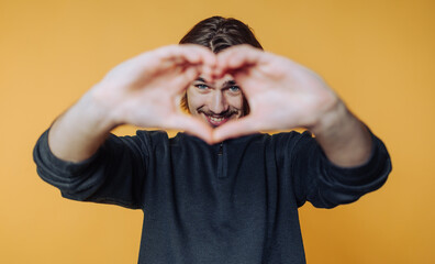 portrait of young man playfully creates a heart shape with his hands on yellow background