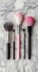 Five makeup brushes of varying sizes and colors arranged on a marble surface; two large powder brushes, two angled liner brushes, and one small detail brush