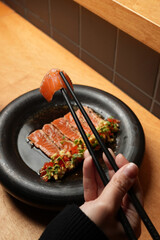 Close-up of a hand with chopsticks picking up a salmon sashimi slice from a gourmet Asian dish