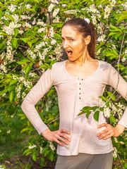 Young woman sneezes in the park against the background of a flowering tree. Allergy to pollen concept.
