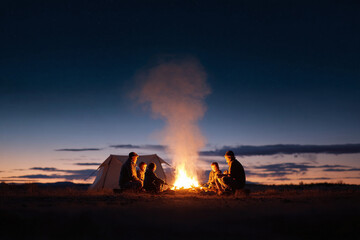 Fototapeta premium Family bonding around a campfire under a starlit sky. Cozy tent glows softly in the background, evoking feelings of togetherness and outdoor adventure. Ideal for travel, lifestyle, family themes.
