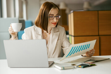 A professional woman meticulously reviews various charts while enjoying a delicious coffee in a stylish workspace