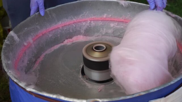 Pink cotton candy being spun in professional cotton candy machine at outdoor event, close up of sugar floss production process with hands in protective gloves
