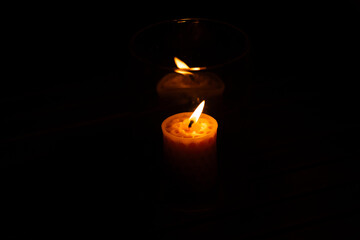 In the darkness, through a glass container, the light of a burning candle reflects, shimmering with soft shades of gold, amber, and gentle rainbow hues.
