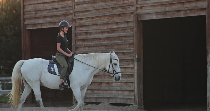 Female equestrian wearing a helmet and casual attire riding a beautiful white horse while leaving the stable at a ranch, enjoying the freedom of the outdoors and equine companionship