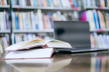 An open book with a stack of books on the table in the library beside a laptop, representing the...