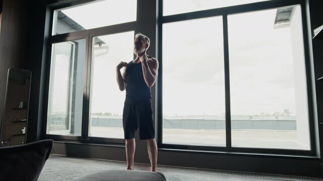 Low angle stab long shot of mature Caucasian muscular man wearing black activewear performing arm circles exercise while standing by panoramic windows in modern minimalist flat