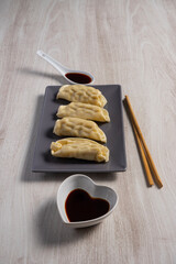Four gyozas on a gray rectangular plate with wooden chopsticks, a spoon, and a bowl of soy sauce on a light wood surface.