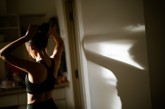 A woman adjusts her hair in a sunlit room, creating a beautiful shadow on the wall, emphasizing her silhouette and the warm ambiance.