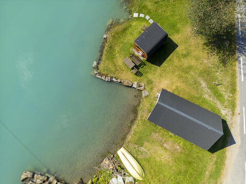 Aerial view of quaint cabins nestled by the turquoise lake's edge, the lush green grass contrasting with the cabins' dark roofs, Oldevatnet, Vestland, Norway.