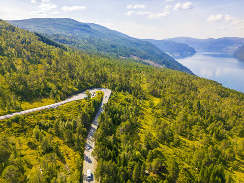 Aerial view of a winding road cutting through lush green forests leading to a tranquil lake surrounded by majestic mountains, Utvik, Vestland, Norway.