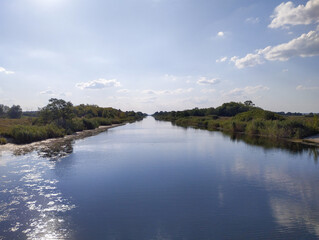 ​A straight canal with a smooth water surface reflecting the blue sky with light white clouds,...