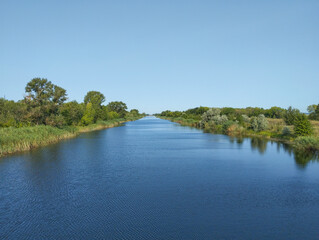 The straight water surface of a canal, covered with a slight ripple, stretches into the distance under a cloudless blue sky.