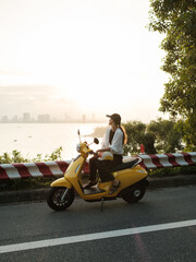 Woman sitting on a yellow scooter by the waterfront road at sunset, enjoying travel and freedom with city skyline in the background.