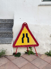 Caution sign indicating road narrowing, positioned against a white wall with textured surface and surrounding greenery, highlighting safety awareness