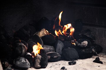Flames dancing atop charcoal in a fireplace, creating a warm glow, surrounded by dark stones, evoking a cozy atmosphere for gatherings and culinary experiences