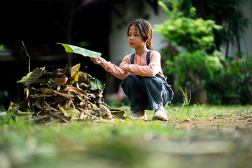 A young Asian girl wearing a pink long-sleeve shirt and denim overalls is crouching in a garden,...