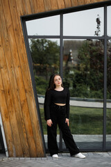 A girl in black trousers and a black blouse stands by a mirrored wall, leaning against it. She has...