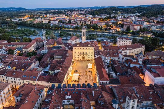 Aerial view of the Palazzo Civico casting a golden glow over a bustling piazza amid the terracotta rooftops and the sinuous Dora Baltea river, Ivrea, Piedmont, Italy.