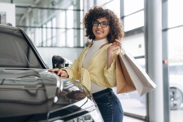 In casual clothes, standing by the vehicle. Pretty woman is in the car retail shop