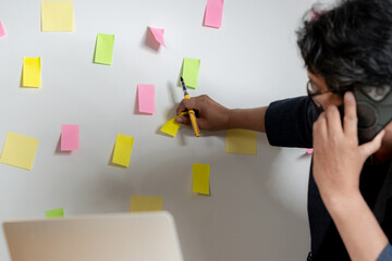 A young businessman writes ideas on sticky notes and attaches them to a wall in his office.