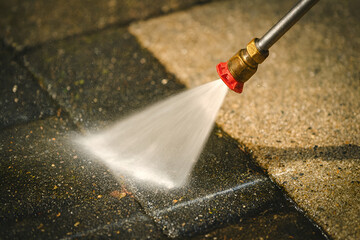 Close-up pressure washer nozzle blasting dirt from patio pavers—clean/dirty contrast on stone. Power washing driveway or walkway; outdoor cleaning, moss and algae removal, home maintenance.