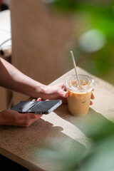 Close-up of a woman holding a smartphone in one hand and a glass of iced coffee in the other,...