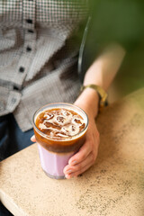 Close-up of a woman's hand holding a glass of iced lavender coffee in a modern coffee shop. Cold...