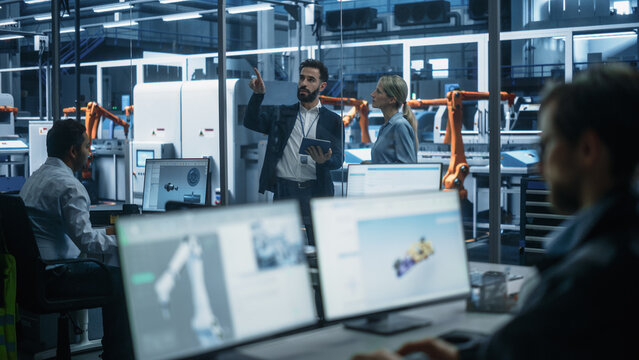 Electric Engine Production At Autonomous Factory With Robotic Arms: Diverse Technicians And Engineers Using Desktop Computers To Control Assembly Line While Managers With Tablet Discussing Project